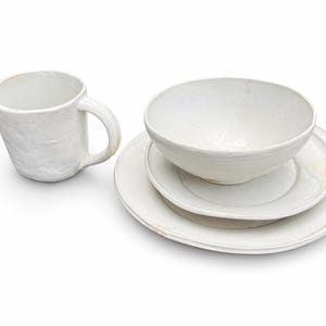 May include: A white ceramic mug, bowl, and two plates set against a white background. The mug has a handle and a textured surface. The bowl sits atop a plate, with another plate beneath. The tableware is likely handmade.