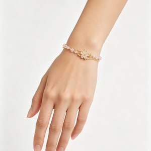 May include: A delicate gold-toned bracelet featuring a central flower-shaped charm with white petals and a series of small, round, pale pink beads strung along a thin gold chain that wraps around the wrist to create a layered appearance.