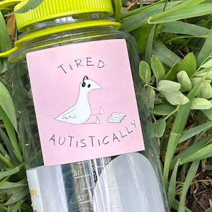 May include: A clear plastic water bottle with a bright lime green screw-top lid. A rectangular pink sticker on the front features a black line drawing of a bird wearing headphones next to a book, with the text TIRED AUTISTICALLY printed.