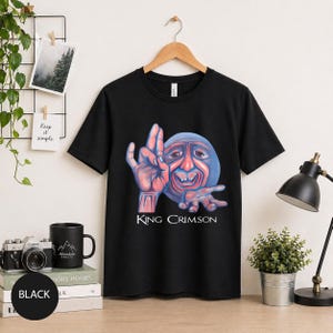 Può includere: T-shirt nera con una grafica di un volto blu con mani rosse e la scritta "King Crimson" in bianco. Il disegno è centrato sulla maglietta. La maglietta è appesa a una gruccia di legno.