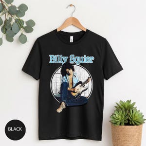 May include: Black t-shirt featuring a graphic of Billy Squier playing a guitar, set within a white circle. The text "Billy Squier" is above the image in a light blue font. The shirt is hanging on a wooden hanger.