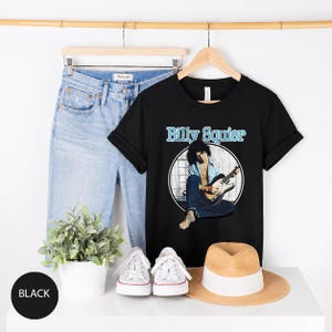 May include: Black t-shirt with the text "Billy Squier" in light blue. The shirt features a graphic of a musician playing a guitar. Light blue jeans, white sneakers, and a straw hat are also visible.