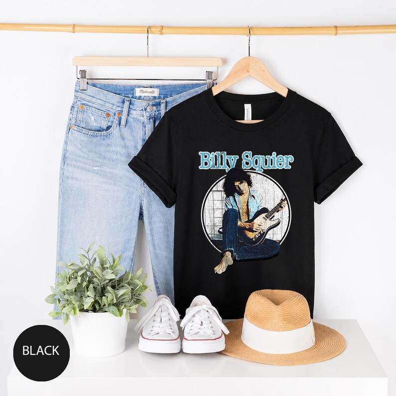 May include: Black t-shirt with the text "Billy Squier" in light blue. The shirt features a graphic of a musician playing a guitar. Light blue jeans, white sneakers, and a straw hat are also visible.