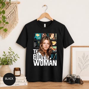 Puede incluir: Camiseta negra con la frase blanca "THE BIONIC WOMAN" y un retrato de mujer. El diseño incluye un collage de imágenes y diagramas. La camiseta está colgada en una percha de madera.