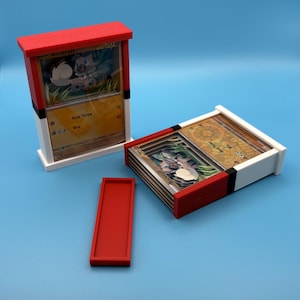 May include: Two rectangular card storage cases designed with a red, white, and black color scheme resembling a Poké Ball. One case stands vertically holding a Rockruff trading card, while the other lies horizontally with a stack of cards inside.