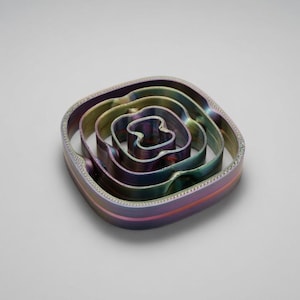 May include: A set of three nested, square-shaped cookie cutters with rounded corners. The cutters feature an iridescent, metallic finish that shifts between shades of purple, blue, and green, with a textured, beaded pattern along the top edges.
