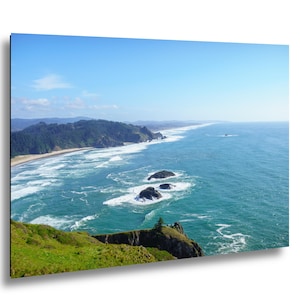 May include: A scenic landscape photograph featuring a coastal view under a clear blue sky. The image captures the ocean meeting the shore, with waves crashing against the rocks. Green vegetation covers the foreground, and a distant shoreline is visible.