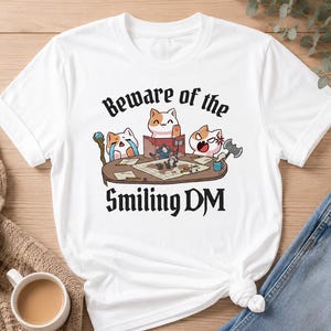 May include: White t-shirt featuring a cartoon illustration of three cats playing a tabletop game. The text "Beware of the Smiling DM" is printed above and below the illustration in a gothic font. The cats are depicted with various expressions.