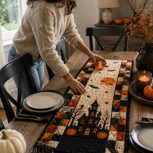 May include: A rectangular quilted table runner featuring a Halloween theme with a central haunted house, bare trees, pumpkins, a full moon, and flying bats. The border consists of a patchwork pattern in black, orange, and cream fabric squares.