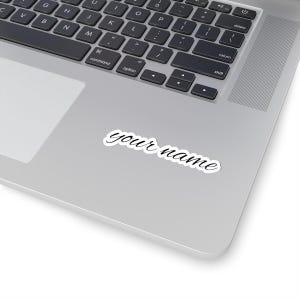 Könnte beinhalten: Ein weißer Aufkleber mit den Worten "your name" in einer kursiven Schriftart. Der Aufkleber befindet sich auf einem silbernen Laptop, wobei die Tastatur im Hintergrund sichtbar ist.