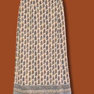 May include: A floor-length skirt in a light beige hue featuring a repeating pattern of small blue and brown paisley-like motifs. The garment includes a wide, decorative border at the hem with intricate blue and brown detailing and two buttons.