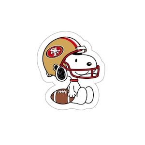 May include: A die-cut sticker featuring a white cartoon dog wearing a gold and red football helmet with the San Francisco 49ers logo on the side. The dog is sitting down and holding a brown football with white laces in its lap.