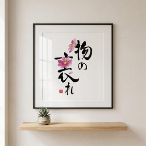 Puede incluir: Impresión artística enmarcada con caligrafía japonesa y flores de acuarela. La obra muestra trazos negros de pincel de caracteres japoneses con flores de acuarela rosas y moradas. Una pequeña planta suculenta en maceta se encuentra en una estantería de madera.