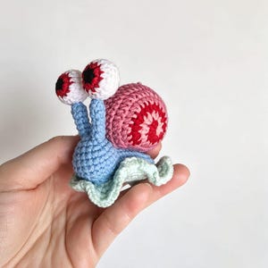 May include: A small, handmade crochet snail toy featuring a light blue body, a ruffled pale green base, and a pink and red spiral shell. The snail has two tall stalks topped with white, round eyes that have prominent red irises and black pupils.
