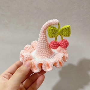 Op de afbeelding: Een handgemaakt gehaakt haaraccessoire in de vorm van een roze en witte hoed met een gerimpelde rand. De hoed is versierd met een groene strik en twee roze kersen. Het accessoire wordt in een hand gehouden.