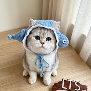 以下が含まれることがあります： 魚の形をした水色のクロシェ編みのペット用帽子。白いスカラップの縁取り、側面のヒレ、結び紐が付いています。グレーと白のトラ猫が着用しています。近くの木製看板にはLTSとLoopThreadStudioの文字が表示されています。