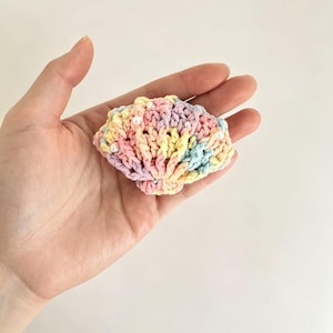 May include: A small, fan-shaped crochet seashell ornament crafted from variegated pastel yarn in shades of pink, yellow, blue, and lavender. The textured, ribbed surface is embellished with several tiny, white, round beads scattered throughout the stitches.