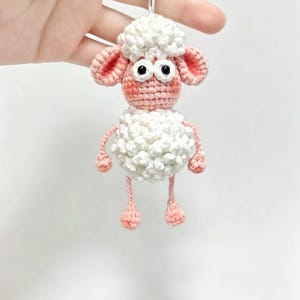 May include: A crochet keychain featuring a small sheep with a white textured wool body and head, pink ears, face, arms, and legs. The sheep has two large white eyes with black pupils and is attached to a silver metal chain and ring for hanging.