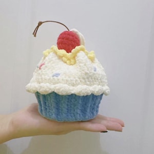 Op de afbeelding: Een gehaakte cupcake met een blauwe basis en witte glazuur. De glazuur is versierd met hagelslag en bekroond met een rode kers. Een bruin touwtje is aan de kers bevestigd.