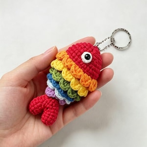 Può includere: Un portachiavi a forma di pesce all'uncinetto con testa rossa, un occhio bianco e nero e squame a strati nei colori arancione, giallo, verde, blu, bianco e viola, che termina con una coda rossa. Un anello in metallo argentato è fissato in cima.