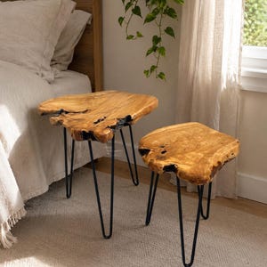 May include: A set of two side tables featuring natural live-edge wood slabs with a polished finish and dark, charred-looking edges. Each table is supported by three slender, black metal hairpin legs that provide a stable base for the wood tops.