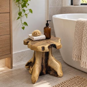 May include: A natural wood stool crafted from a tree stump with a smooth, circular top and a sturdy, organic base featuring dark, charred-looking accents. The stool is topped with a folded white waffle-weave cloth, a natural sponge, and a brown bottle.