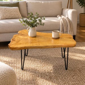 May include: A live edge coffee table featuring a natural light brown wood slab top with an irregular organic shape, supported by four thin black metal hairpin legs. A white ceramic pitcher with green foliage and a ceramic mug sit on the surface.