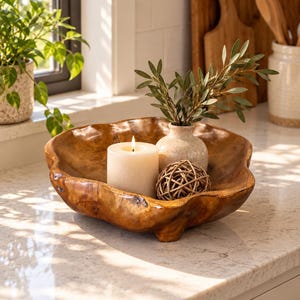 May include: A large, organic-shaped wooden bowl with a polished finish containing a lit white pillar candle, a small speckled ceramic vase with green leafy stems, and a decorative woven rattan sphere resting inside the bowl's curved basin.