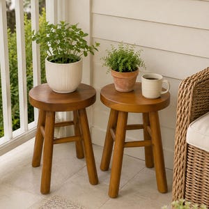 May include: Two round wooden stools with four legs each. One stool holds a white ribbed ceramic pot with a green plant. The other stool holds a terracotta pot with a green plant and a beige ceramic mug filled with a dark liquid beverage.