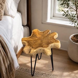 May include: A side table featuring a natural, irregular-shaped light brown wood slab top with visible grain patterns. The tabletop is supported by three thin, black metal hairpin legs that provide a stable base for the organic wood surface.