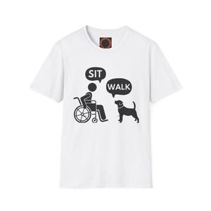 Puede incluir: Camiseta blanca con un gráfico negro de una persona en silla de ruedas y un perro. Las burbujas de diálogo dicen "SIT" y "WALK". La camiseta tiene cuello redondo y mangas cortas.
