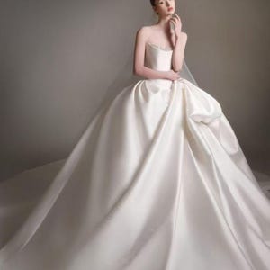 May include: A floor-length, strapless ivory wedding gown featuring a structured bodice with subtle beaded detailing along the neckline and a voluminous, full-length skirt made of smooth, satin-like fabric that drapes into a wide, sweeping train.