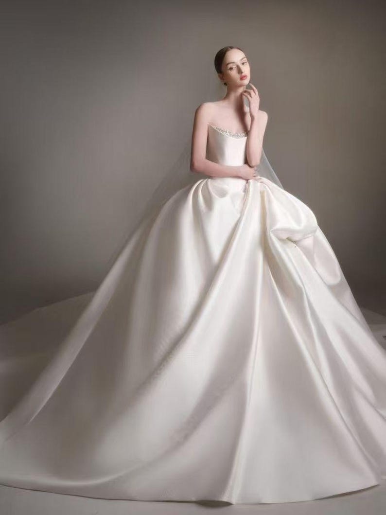 May include: A floor-length, strapless ivory wedding gown featuring a structured bodice with subtle beaded detailing along the neckline and a voluminous, full-length skirt made of smooth, satin-like fabric that drapes into a wide, sweeping train.