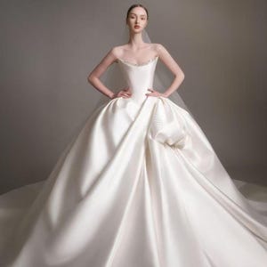 May include: A strapless white ball gown with a structured bodice featuring a subtle beaded neckline and a voluminous, floor-length skirt with deep folds. The dress is paired with a sheer, lightweight veil that drapes over the shoulders.