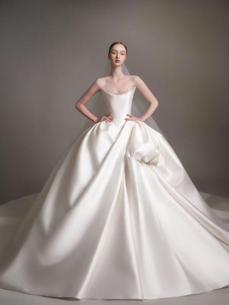 May include: A strapless white ball gown with a structured bodice featuring a subtle beaded neckline and a voluminous, floor-length skirt with deep folds. The dress is paired with a sheer, lightweight veil that drapes over the shoulders.