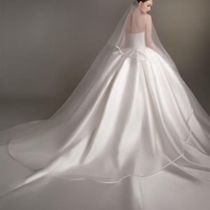May include: A floor-length white satin ball gown with a strapless bodice and a voluminous skirt that extends into a long train. A sheer, translucent veil is draped over the back, extending from the head down to the floor over the dress.