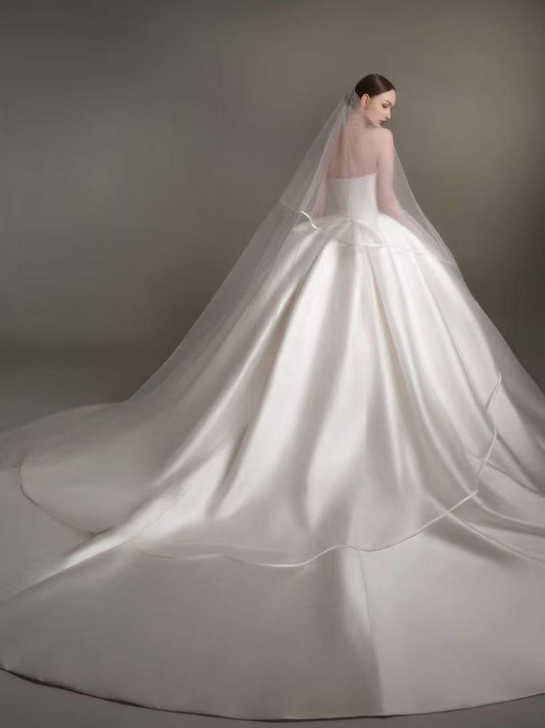 May include: A floor-length white satin ball gown with a strapless bodice and a voluminous skirt that extends into a long train. A sheer, translucent veil is draped over the back, extending from the head down to the floor over the dress.