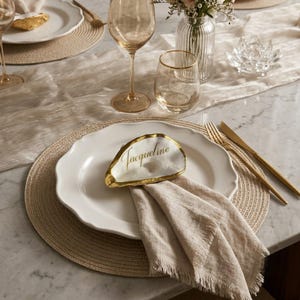 May include: A white scalloped ceramic dinner plate featuring a natural oyster shell place card with gold-painted edges and the name Jacqueline written in gold script. A beige linen napkin with frayed edges is folded underneath the shell.