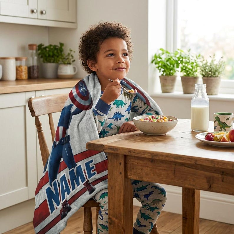 May include: A child wearing dinosaur pajamas and a Captain America blanket sits at a wooden table eating cereal. The blanket features a Captain America design with the name "NAME" in large letters. A bottle of milk and a dinosaur mug are on the table.