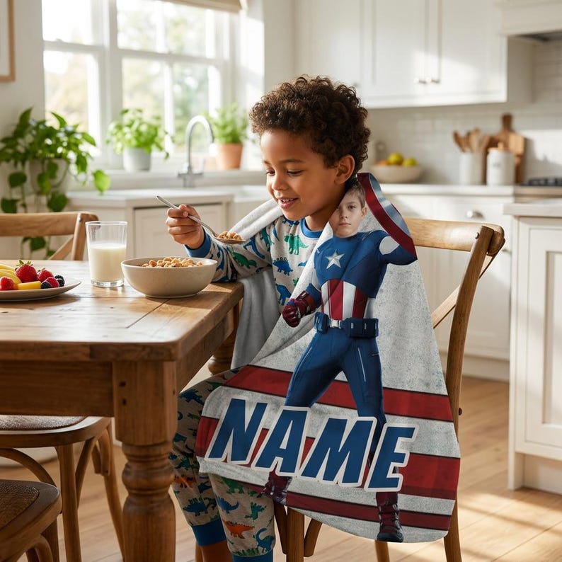 May include: A Captain America-themed blanket with a personalized name banner. The blanket features a graphic of Captain America in his iconic blue, red, and white suit. The blanket is draped over a child, who is sitting at a wooden table.