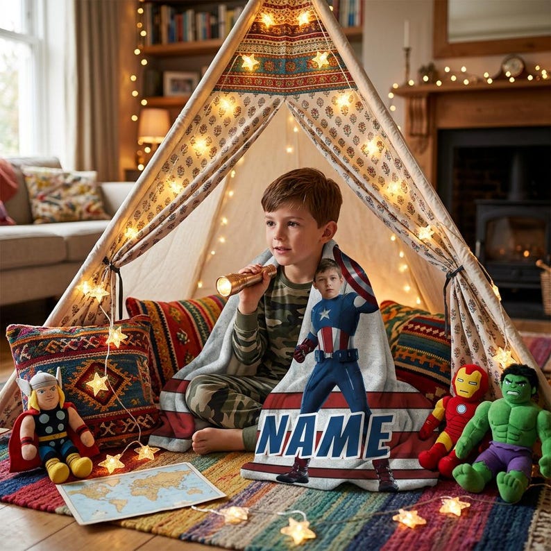 May include: A child sits inside a decorated teepee, holding a flashlight and wrapped in a Captain America blanket. The blanket has the word "NAME" on it. Plush superhero toys and a world map are nearby, with string lights adding a warm glow.