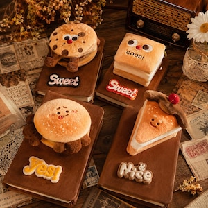 May include: Four brown notebooks featuring plush food-themed covers: a chocolate chip cookie, a square sandwich, a round burger, and a slice of pie. Each notebook has a decorative patch with the text Sweet, Sweet, Yes!, or Nice attached.