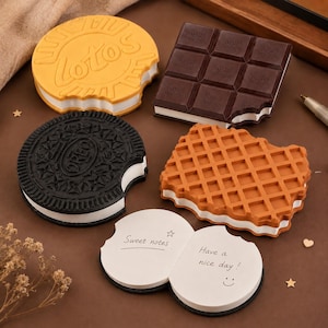 Op de afbeelding: Een verzameling notitieblokken in de vorm van voedsel, waaronder een geel rond Lotos-koekje, een donkerbruine vierkante chocoladereep, een zwart rond Oreo-koekje en een beige rechthoekige wafel, plus een open rond notitieblok met de tekst Sweet notes Have a nice day.