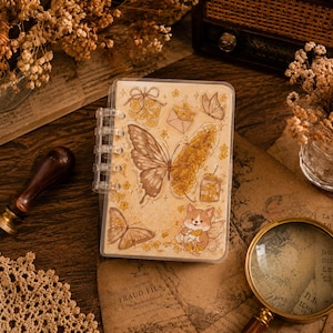 Op de afbeelding: Een ringbandnotitieboek van doorzichtig plastic met een crèmekleurige kaft, voorzien van illustraties van een grote vlinder met één vleugel van gele bloemen, een kleinere vlinder, een corgi, een envelop en een potje in bruine en gele tinten.