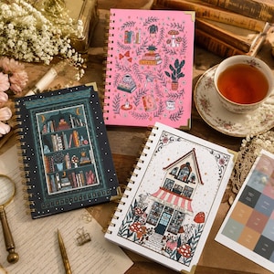 Può includere: Tre agende a spirale con copertine illustrate. Una è rosa con motivi botanici e di libri, una blu scuro con l'illustrazione di una libreria e una bianca che raffigura un edificio con il testo The Reading Planner.