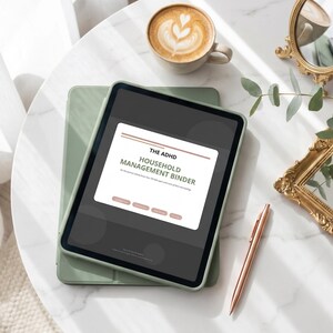 Op de afbeelding: Een digitale tablet met de tekst "THE ADHD HOUSEHOLD MANAGEMENT BINDER" op het scherm. De tablet ligt op een groene hoes, naast een kop koffie met latte art, een gouden spiegel, een pen en een plant op een wit marmeren oppervlak.