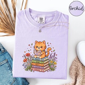 May include: A light purple t-shirt featuring a graphic of an orange cat wearing a rainbow scarf and reading a book labelled Love. The cat sits on a stack of five books labelled Peace, Kind, Unity, Justice, and Change, surrounded by colourful flowers.