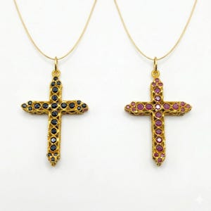 Puede incluir: Dos colgantes de cruz dorados con adornos de piedras preciosas cuelgan de delicadas cadenas doradas. Una cruz presenta piedras preciosas de color azul oscuro, mientras que la otra muestra piedras preciosas rojas. Las cruces tienen detalles ornamentados.