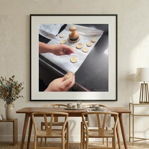 May include: A framed image shows a close-up of a person's hands working with round cookies and a wooden stamp. The cookies are on parchment paper. A wooden stamp is in the center of the image. The cookies have a design stamped on them.