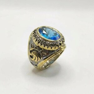 May include: A gold and silver-toned ring with a large, oval, light blue gemstone. The ring features the word "BISHOP" around the gemstone and a crest on the side. The ring is likely a class ring or a commemorative piece.